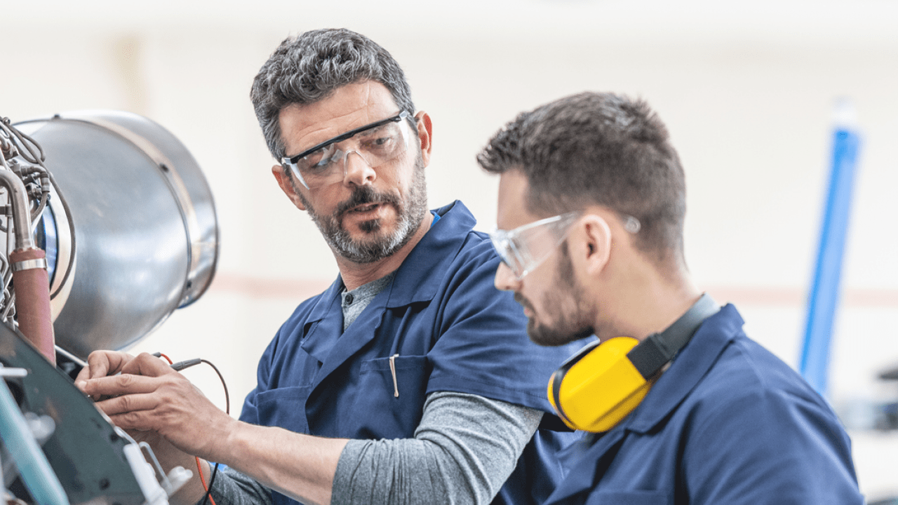 Young aviation mechanics studying to repair engine medium shot. Helicopter hangar workers in protective equipment examining jet, side view